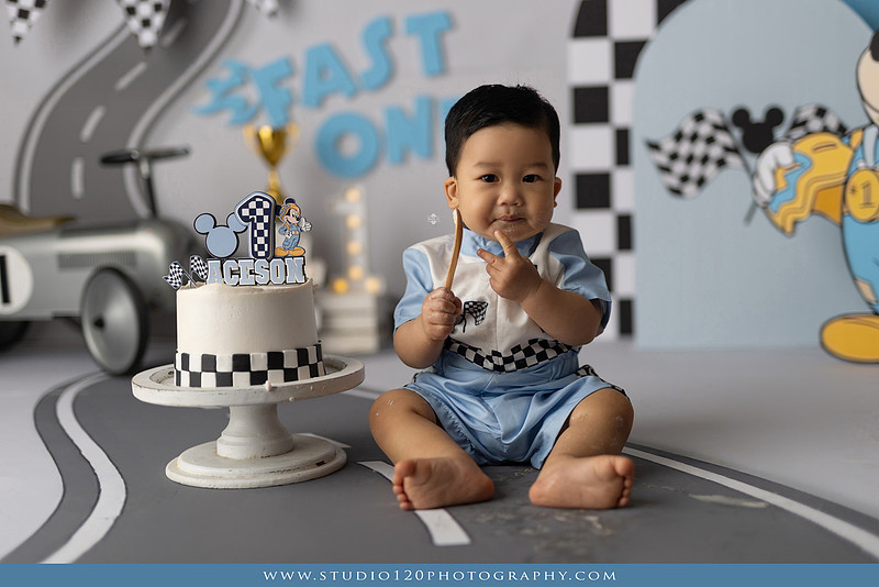 Fast One Mickey Mouse race car cake smash featuring custom race track backdrop, checkered flags, vintage race car prop, and first birthday cake in San Diego