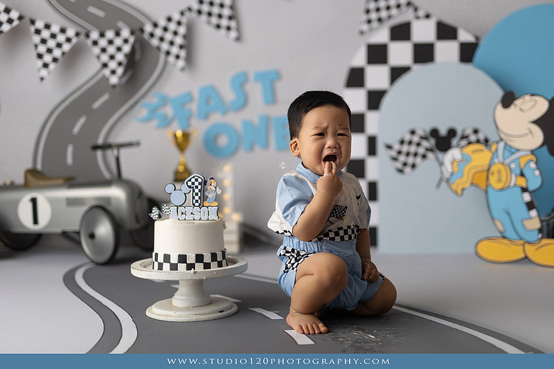 Fast One Mickey Mouse race car cake smash featuring custom race track backdrop, checkered flags, vintage race car prop, and first birthday cake in San Diego