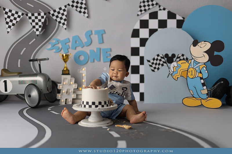 Fast One Mickey Mouse race car cake smash featuring custom race track backdrop, checkered flags, vintage race car prop, and first birthday cake in San Diego