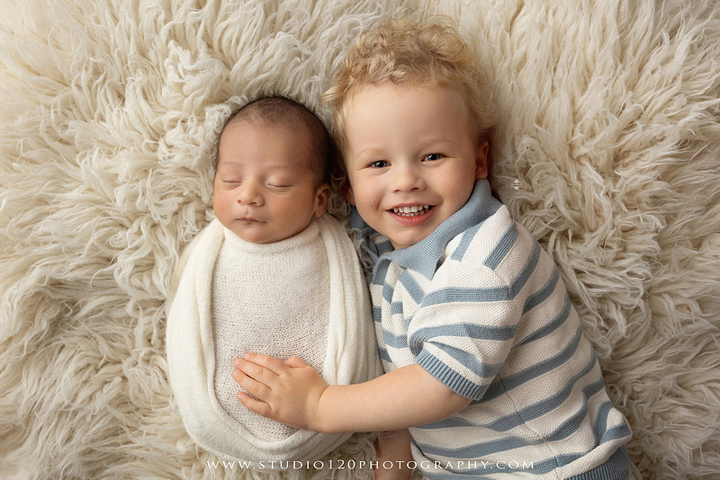 Big brother smiling beside his newborn baby brother during a sibling newborn photography session.