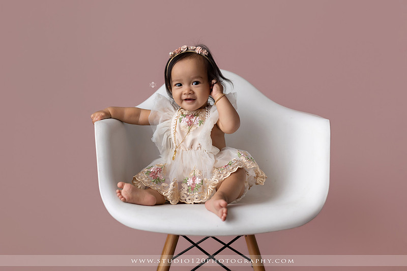 One-year-old girl wearing a floral dress sitting in a modern white chair against a blush pink backdrop during a first birthday milestone portrait session at Studio 120 Photography in Ramona, California.