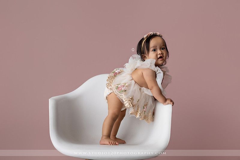 One-year-old girl wearing a floral dress sitting in a modern white chair against a blush pink backdrop during a first birthday milestone portrait session at Studio 120 Photography in Ramona, California.