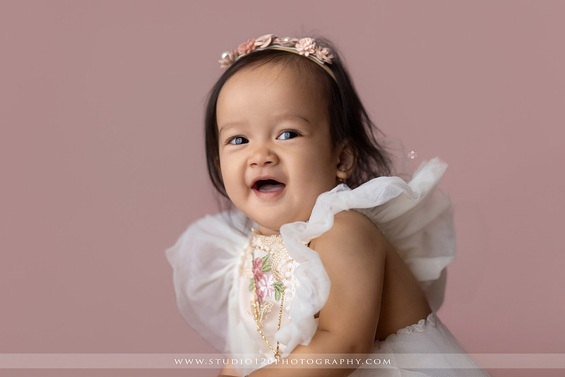 One-year-old girl wearing a floral dress sitting in a modern white chair against a blush pink backdrop during a first birthday milestone portrait session at Studio 120 Photography in Ramona, California.