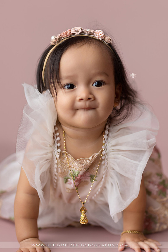 One-year-old girl wearing a floral dress sitting in a modern white chair against a blush pink backdrop during a first birthday milestone portrait session at Studio 120 Photography in Ramona, California.