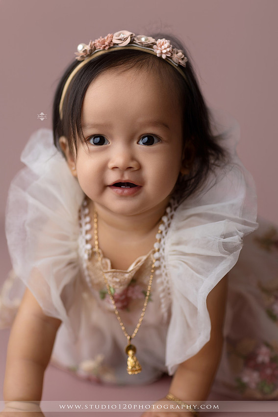One-year-old girl wearing a floral dress sitting in a modern white chair against a blush pink backdrop during a first birthday milestone portrait session at Studio 120 Photography in Ramona, California.