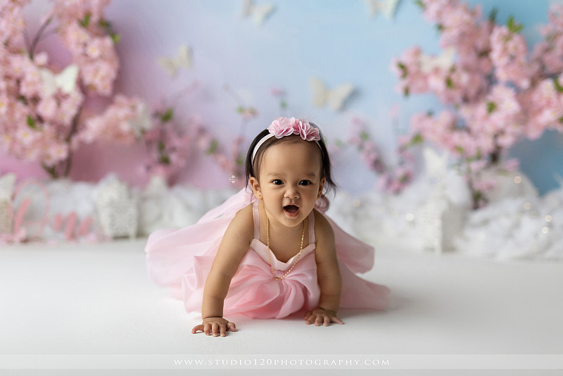 One-year-old girl enjoying an elegant blush pink floral cake smash with cherry blossoms, butterflies, and a custom birthday cake during a luxury first birthday photography session at Studio 120 Photography in Ramona, California.