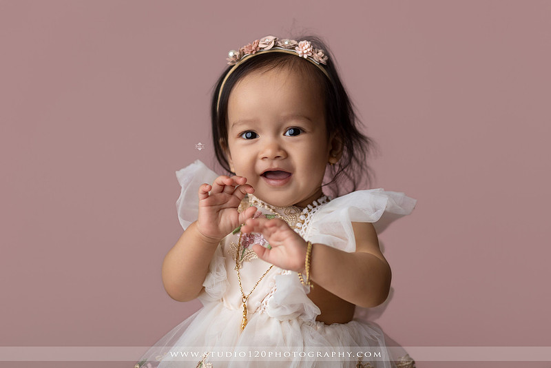 One-year-old girl wearing a floral dress sitting in a modern white chair against a blush pink backdrop during a first birthday milestone portrait session at Studio 120 Photography in Ramona, California.