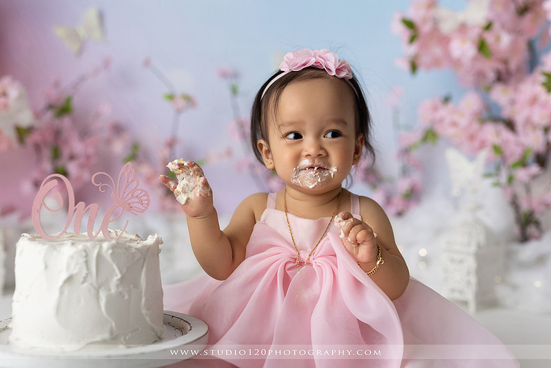 One-year-old girl enjoying an elegant blush pink floral cake smash with cherry blossoms, butterflies, and a custom birthday cake during a luxury first birthday photography session at Studio 120 Photography in Ramona, California.