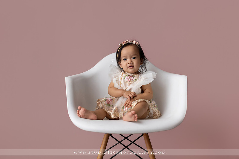 One-year-old girl wearing a floral dress sitting in a modern white chair against a blush pink backdrop during a first birthday milestone portrait session at Studio 120 Photography in Ramona, California.