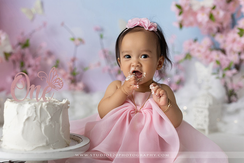One-year-old girl enjoying an elegant blush pink floral cake smash with cherry blossoms, butterflies, and a custom birthday cake during a luxury first birthday photography session at Studio 120 Photography in Ramona, California.