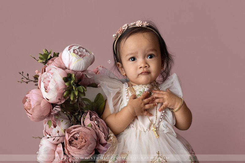 One-year-old girl wearing a floral dress sitting in a modern white chair against a blush pink backdrop during a first birthday milestone portrait session at Studio 120 Photography in Ramona, California.