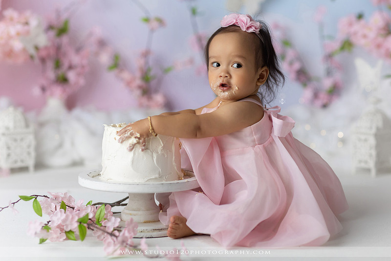 One-year-old girl enjoying an elegant blush pink floral cake smash with cherry blossoms, butterflies, and a custom birthday cake during a luxury first birthday photography session at Studio 120 Photography in Ramona, California.