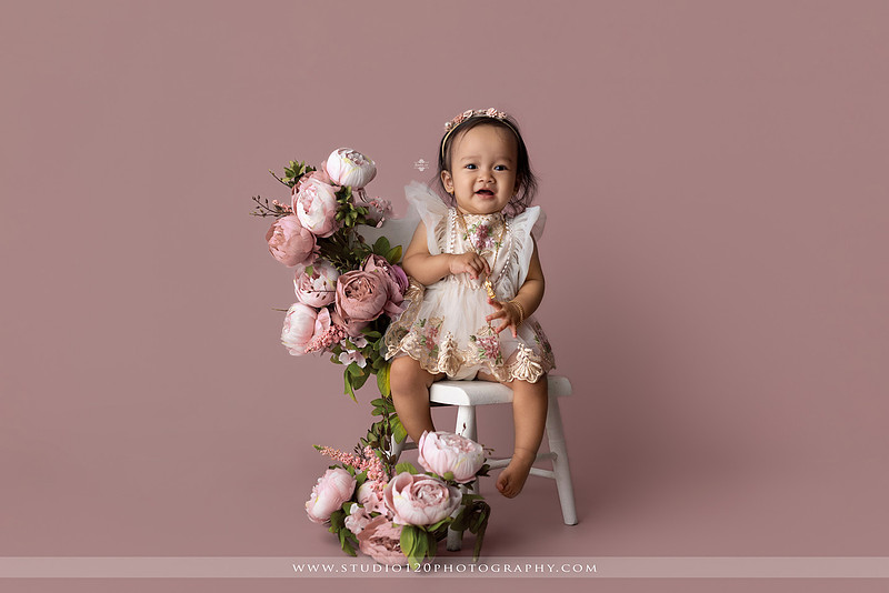 One-year-old girl wearing a floral dress sitting in a modern white chair against a blush pink backdrop during a first birthday milestone portrait session at Studio 120 Photography in Ramona, California.