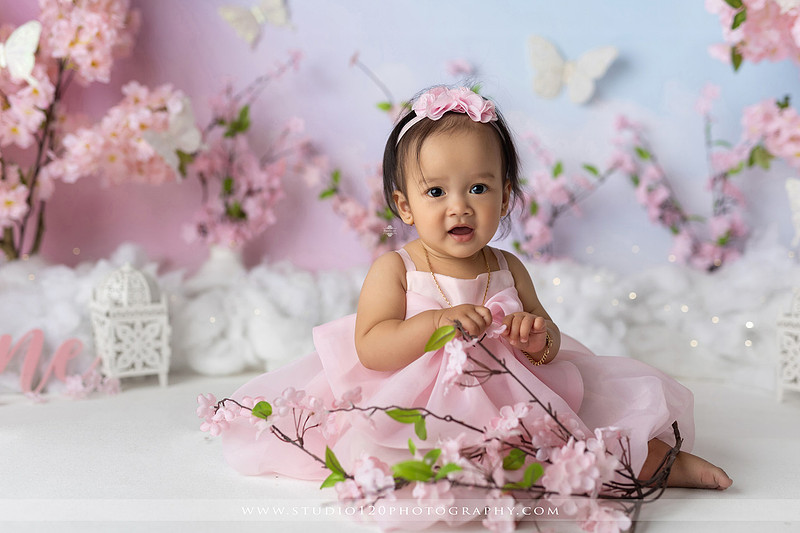 One-year-old girl enjoying an elegant blush pink floral cake smash with cherry blossoms, butterflies, and a custom birthday cake during a luxury first birthday photography session at Studio 120 Photography in Ramona, California.