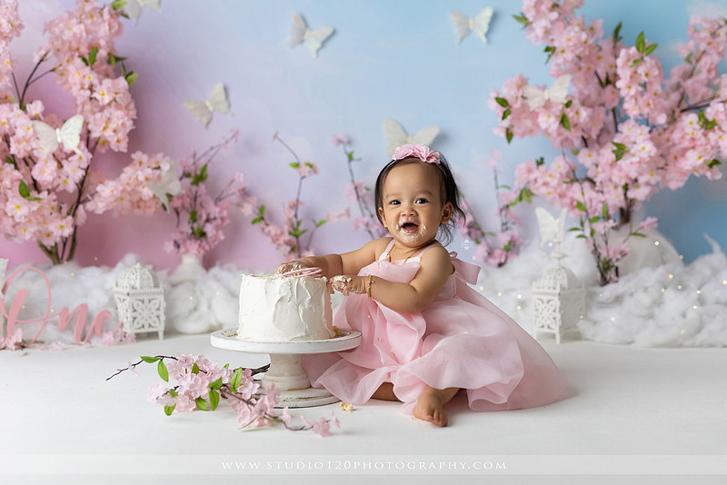 One-year-old girl enjoying an elegant blush pink floral cake smash with cherry blossoms, butterflies, and a custom birthday cake during a luxury first birthday photography session at Studio 120 Photography in Ramona, California.