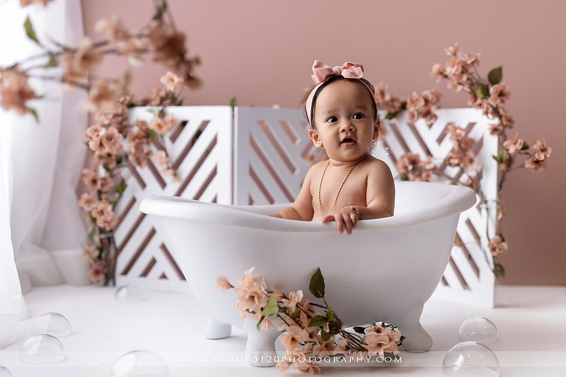 One-year-old girl splashing in a white clawfoot tub surrounded by blush pink flowers during a luxury floral milk bath photography session for her first birthday at Studio 120 Photography in Ramona, California.
