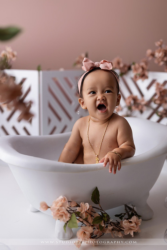 One-year-old girl splashing in a white clawfoot tub surrounded by blush pink flowers during a luxury floral milk bath photography session for her first birthday at Studio 120 Photography in Ramona, California.