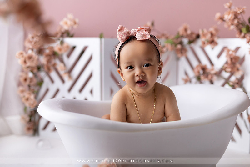 One-year-old girl splashing in a white clawfoot tub surrounded by blush pink flowers during a luxury floral milk bath photography session for her first birthday at Studio 120 Photography in Ramona, California.