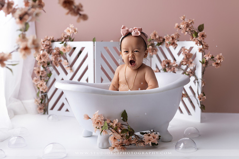 One-year-old girl splashing in a white clawfoot tub surrounded by blush pink flowers during a luxury floral milk bath photography session for her first birthday at Studio 120 Photography in Ramona, California.