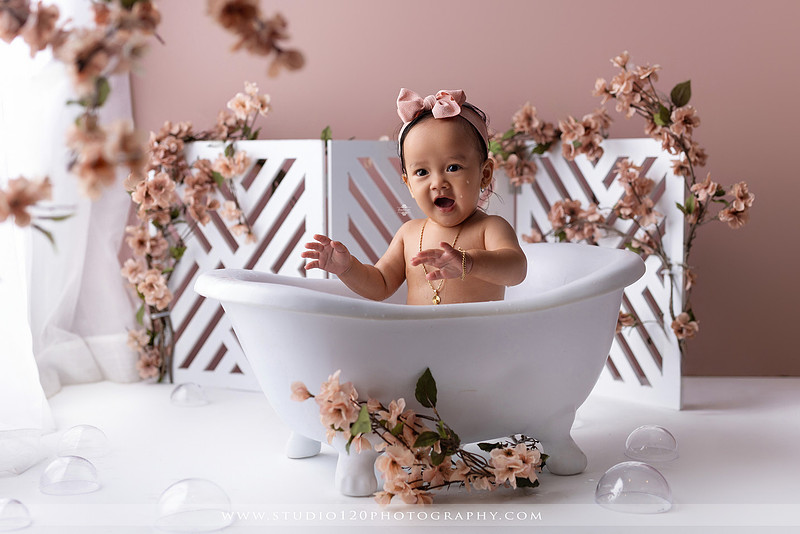 One-year-old girl splashing in a white clawfoot tub surrounded by blush pink flowers during a luxury floral milk bath photography session for her first birthday at Studio 120 Photography in Ramona, California.