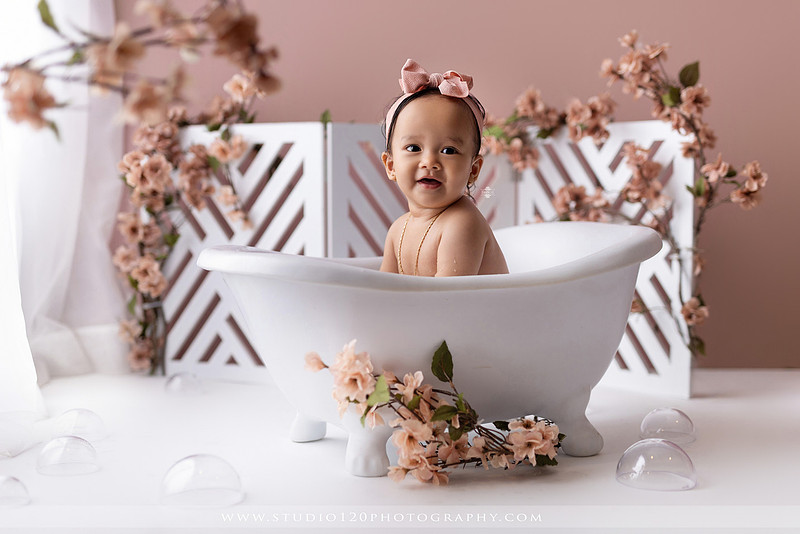 One-year-old girl splashing in a white clawfoot tub surrounded by blush pink flowers during a luxury floral milk bath photography session for her first birthday at Studio 120 Photography in Ramona, California.