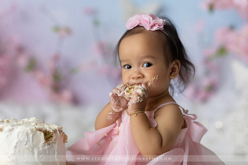 One-year-old girl enjoying an elegant blush pink floral cake smash with cherry blossoms, butterflies, and a custom birthday cake during a luxury first birthday photography session at Studio 120 Photography in Ramona, California.