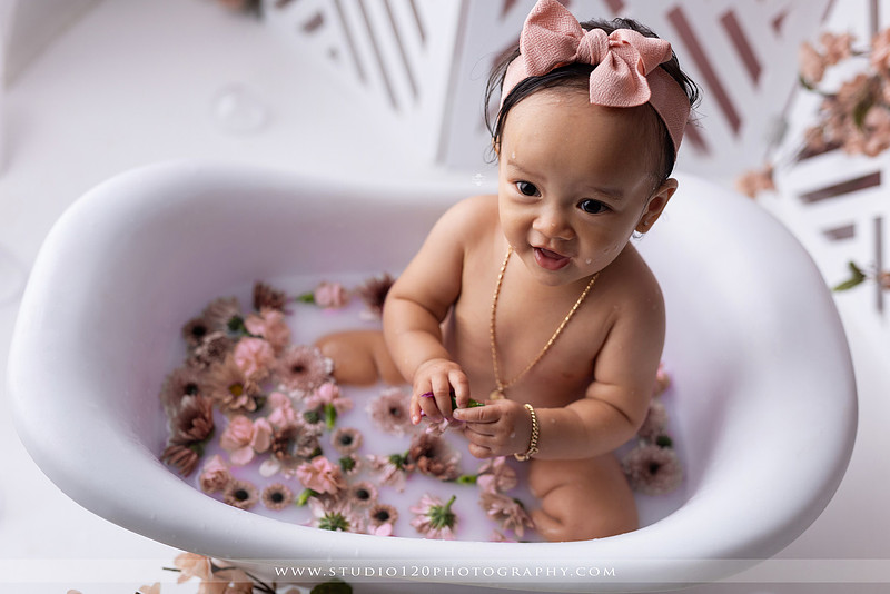 One-year-old girl splashing in a white clawfoot tub surrounded by blush pink flowers during a luxury floral milk bath photography session for her first birthday at Studio 120 Photography in Ramona, California.