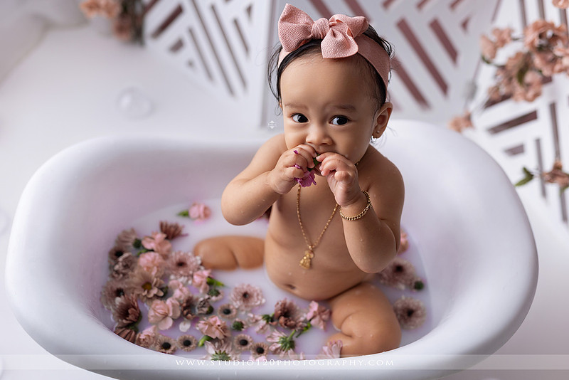 One-year-old girl splashing in a white clawfoot tub surrounded by blush pink flowers during a luxury floral milk bath photography session for her first birthday at Studio 120 Photography in Ramona, California.