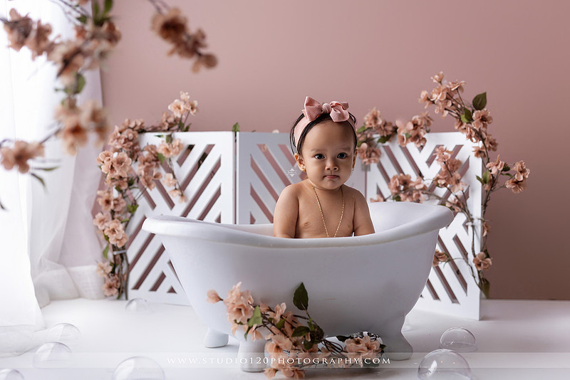 One-year-old girl splashing in a white clawfoot tub surrounded by blush pink flowers during a luxury floral milk bath photography session for her first birthday at Studio 120 Photography in Ramona, California.