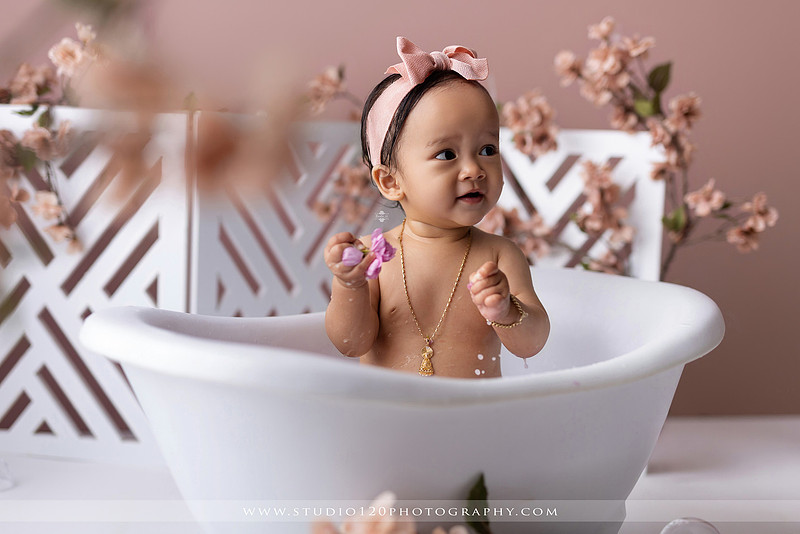 One-year-old girl splashing in a white clawfoot tub surrounded by blush pink flowers during a luxury floral milk bath photography session for her first birthday at Studio 120 Photography in Ramona, California.
