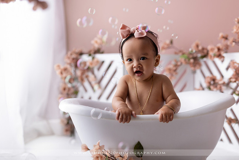One-year-old girl splashing in a white clawfoot tub surrounded by blush pink flowers during a luxury floral milk bath photography session for her first birthday at Studio 120 Photography in Ramona, California.