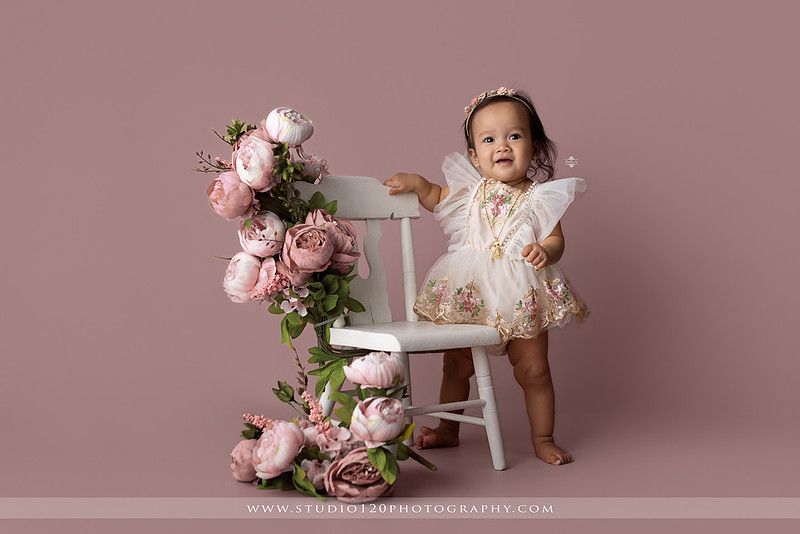 One-year-old girl wearing a floral dress sitting in a modern white chair against a blush pink backdrop during a first birthday milestone portrait session at Studio 120 Photography in Ramona, California.