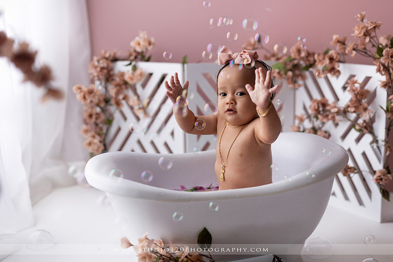 One-year-old girl splashing in a white clawfoot tub surrounded by blush pink flowers during a luxury floral milk bath photography session for her first birthday at Studio 120 Photography in Ramona, California.