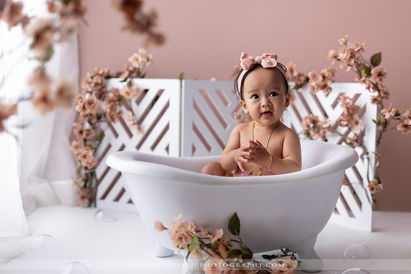 One-year-old girl splashing in a white clawfoot tub surrounded by blush pink flowers during a luxury floral milk bath photography session for her first birthday at Studio 120 Photography in Ramona, California.