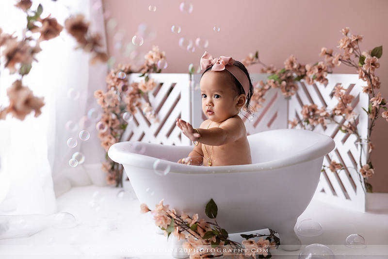One-year-old girl splashing in a white clawfoot tub surrounded by blush pink flowers during a luxury floral milk bath photography session for her first birthday at Studio 120 Photography in Ramona, California.