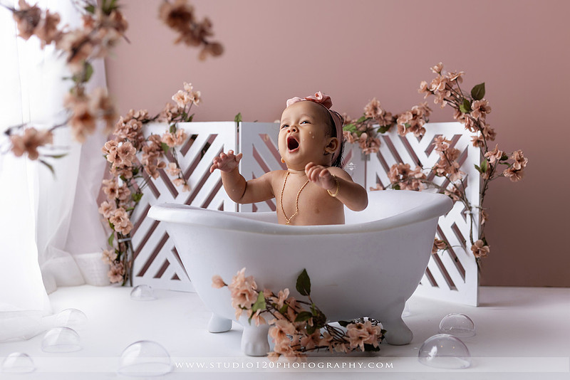 One-year-old girl splashing in a white clawfoot tub surrounded by blush pink flowers during a luxury floral milk bath photography session for her first birthday at Studio 120 Photography in Ramona, California.