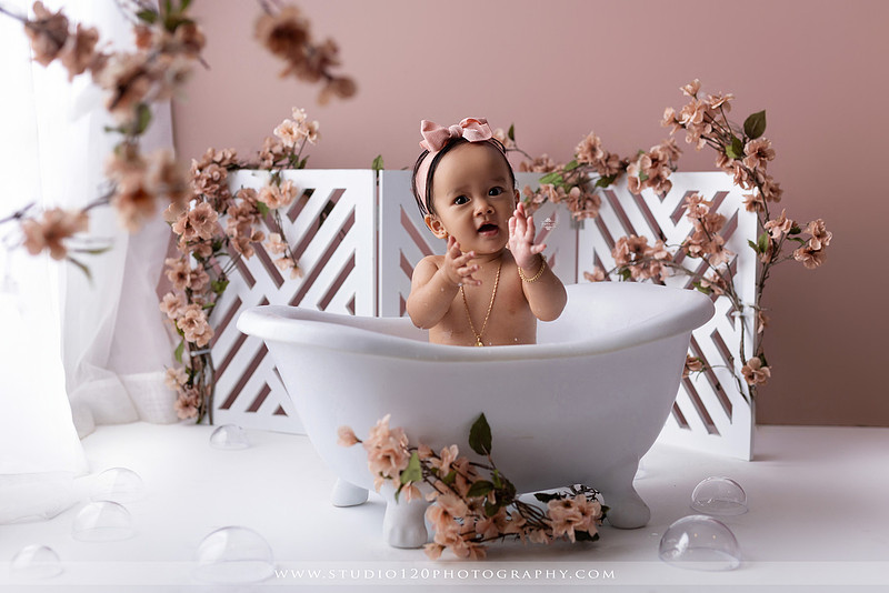 One-year-old girl splashing in a white clawfoot tub surrounded by blush pink flowers during a luxury floral milk bath photography session for her first birthday at Studio 120 Photography in Ramona, California.