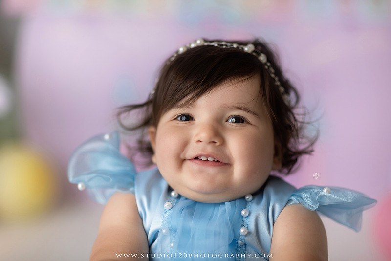 Disneyland-inspired first birthday cake smash featuring a baby girl in a blue princess dress with pastel balloons, whimsical amusement park décor, custom birthday backdrop, milestone portraits, and cake smash photography by Studio 120 Photography in Ramona, serving San Diego County.