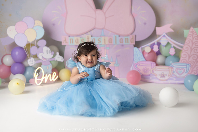 Disneyland-inspired first birthday cake smash featuring a baby girl in a blue princess dress with pastel balloons, whimsical amusement park décor, custom birthday backdrop, milestone portraits, and cake smash photography by Studio 120 Photography in Ramona, serving San Diego County.