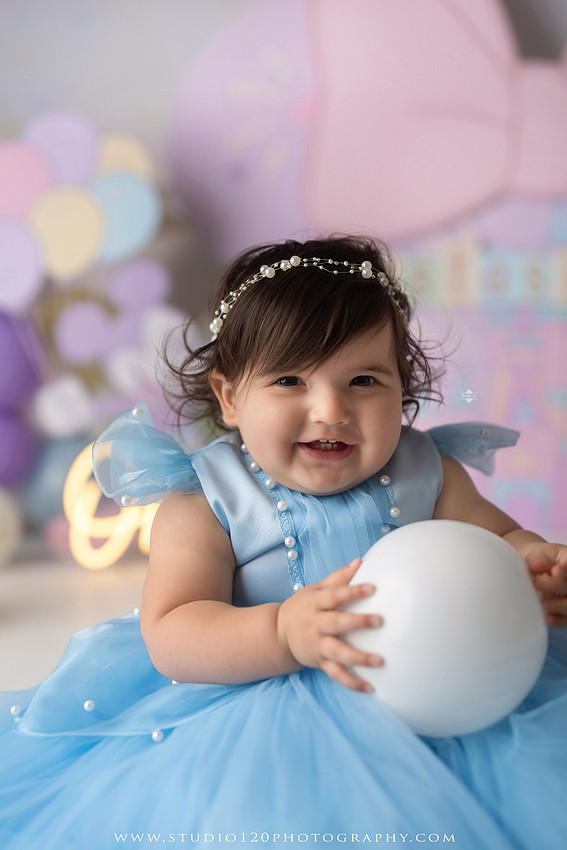 Disneyland-inspired first birthday cake smash featuring a baby girl in a blue princess dress with pastel balloons, whimsical amusement park décor, custom birthday backdrop, milestone portraits, and cake smash photography by Studio 120 Photography in Ramona, serving San Diego County.