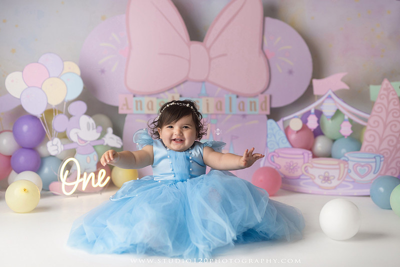 Disneyland-inspired first birthday cake smash featuring a baby girl in a blue princess dress with pastel balloons, whimsical amusement park décor, custom birthday backdrop, milestone portraits, and cake smash photography by Studio 120 Photography in Ramona, serving San Diego County.