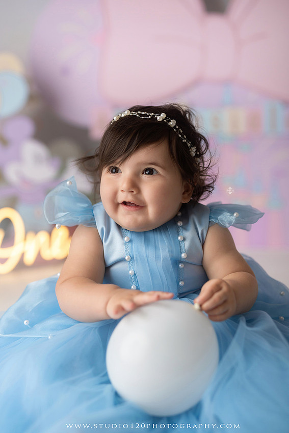 Disneyland-inspired first birthday cake smash featuring a baby girl in a blue princess dress with pastel balloons, whimsical amusement park décor, custom birthday backdrop, milestone portraits, and cake smash photography by Studio 120 Photography in Ramona, serving San Diego County.