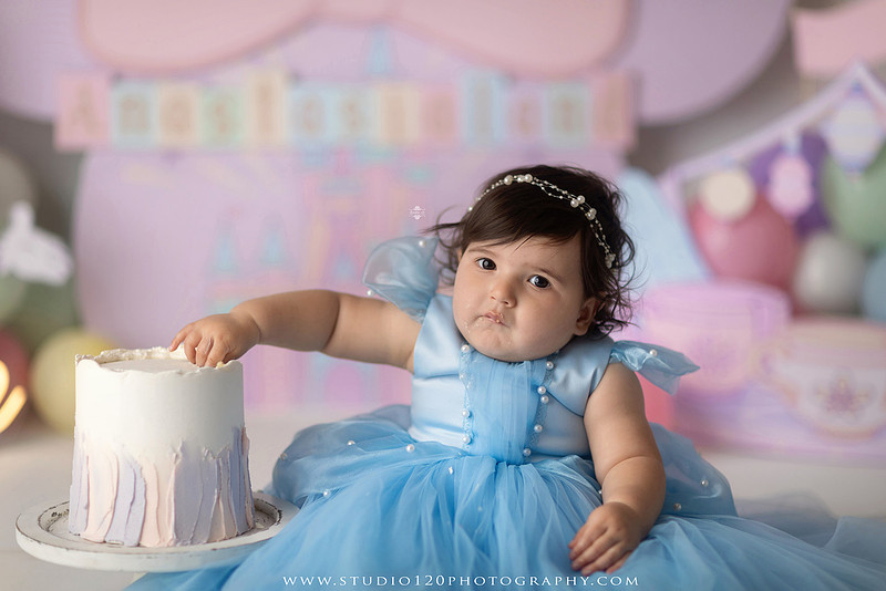 Disneyland-inspired first birthday cake smash featuring a baby girl in a blue princess dress with pastel balloons, whimsical amusement park décor, custom birthday backdrop, milestone portraits, and cake smash photography by Studio 120 Photography in Ramona, serving San Diego County.