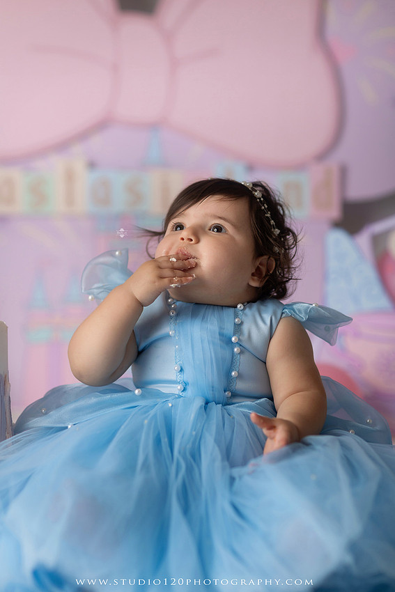 Disneyland-inspired first birthday cake smash featuring a baby girl in a blue princess dress with pastel balloons, whimsical amusement park décor, custom birthday backdrop, milestone portraits, and cake smash photography by Studio 120 Photography in Ramona, serving San Diego County.