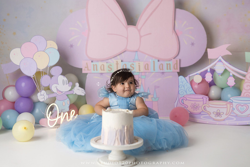 Disneyland-inspired first birthday cake smash featuring a baby girl in a blue princess dress with pastel balloons, whimsical amusement park décor, custom birthday backdrop, milestone portraits, and cake smash photography by Studio 120 Photography in Ramona, serving San Diego County.