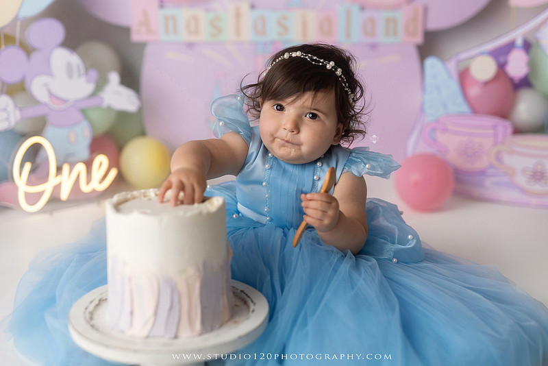 Disneyland-inspired first birthday cake smash featuring a baby girl in a blue princess dress with pastel balloons, whimsical amusement park décor, custom birthday backdrop, milestone portraits, and cake smash photography by Studio 120 Photography in Ramona, serving San Diego County.