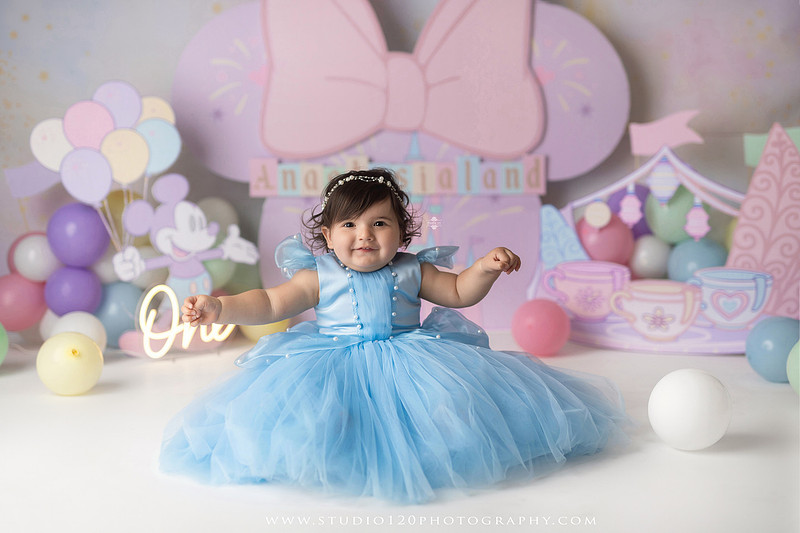 Disneyland-inspired first birthday cake smash featuring a baby girl in a blue princess dress with pastel balloons, whimsical amusement park décor, custom birthday backdrop, milestone portraits, and cake smash photography by Studio 120 Photography in Ramona, serving San Diego County.