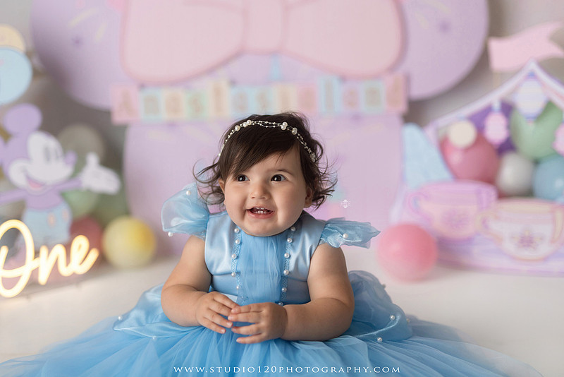Disneyland-inspired first birthday cake smash featuring a baby girl in a blue princess dress with pastel balloons, whimsical amusement park décor, custom birthday backdrop, milestone portraits, and cake smash photography by Studio 120 Photography in Ramona, serving San Diego County.
