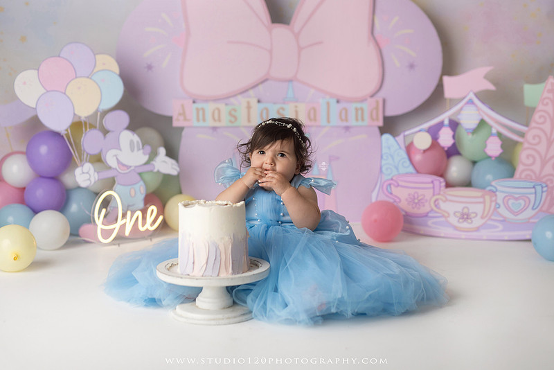 Disneyland-inspired first birthday cake smash featuring a baby girl in a blue princess dress with pastel balloons, whimsical amusement park décor, custom birthday backdrop, milestone portraits, and cake smash photography by Studio 120 Photography in Ramona, serving San Diego County.