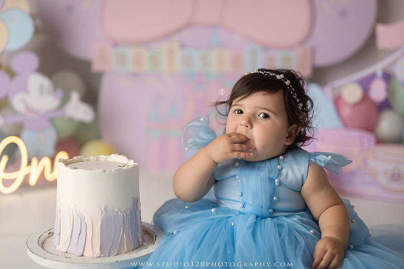 Disneyland-inspired first birthday cake smash featuring a baby girl in a blue princess dress with pastel balloons, whimsical amusement park décor, custom birthday backdrop, milestone portraits, and cake smash photography by Studio 120 Photography in Ramona, serving San Diego County.